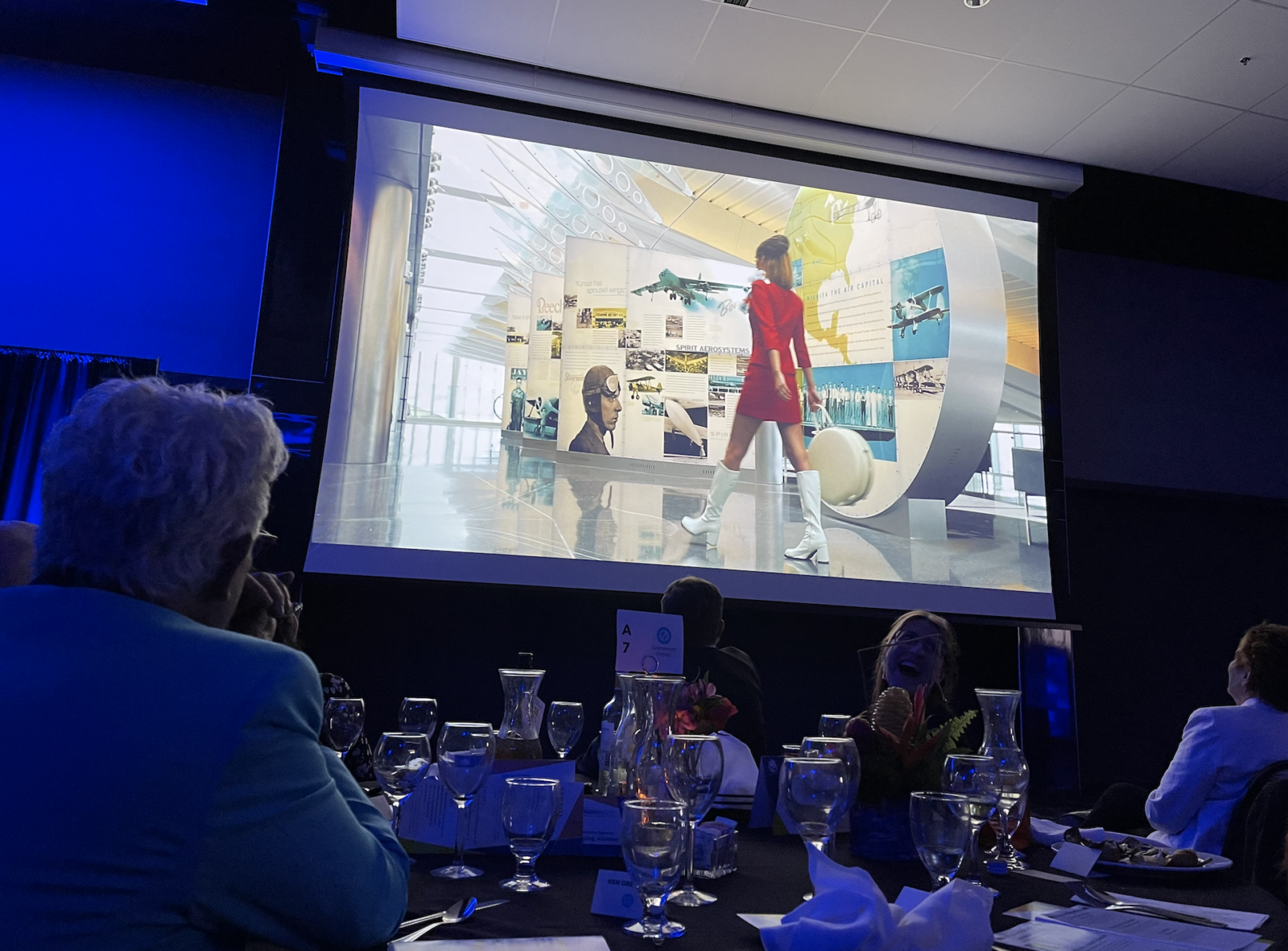 Image of GiGi, Greteman Group's mascot walking through the airport projected onto a screen for a ballroom full of the Chamber's Honors Night attendees to see as Greteman Group received the Over the Years Awars